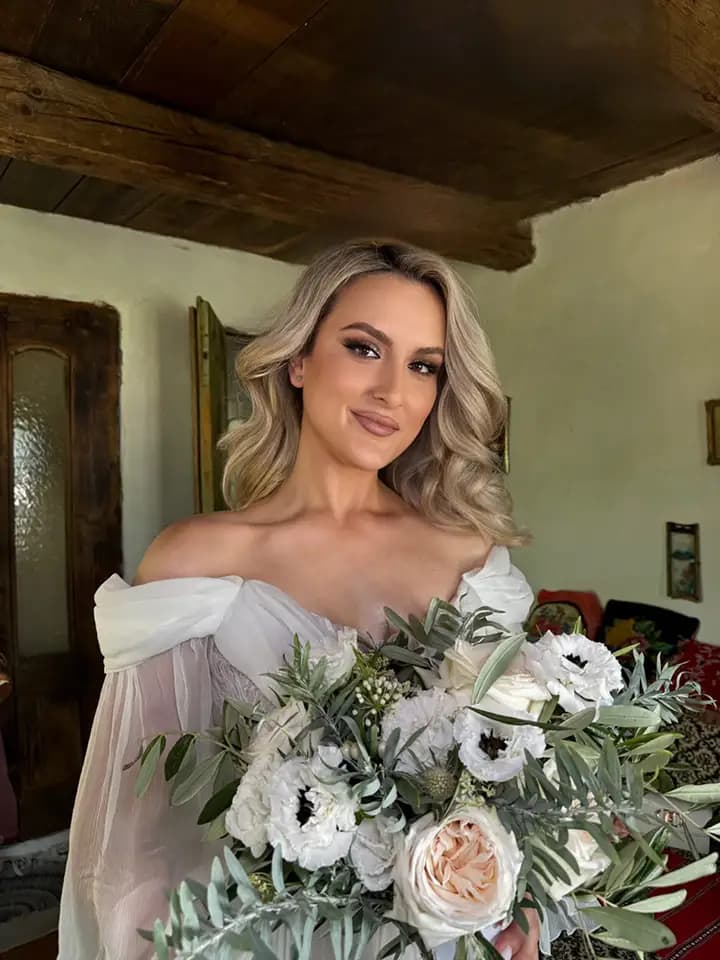 Mireasă cu buchet de anemone albe, trandafiri de grădină și frunze de măslin, într-o cameră rustică